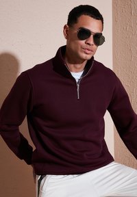 Sudadera burdeos con media cremallera, fabricada en un tejido suave, que presenta un cuello alto y una cremallera elegante. Combinada con pantalones blancos y gafas de sol oscuras.
