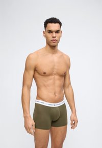 Άνδρας χωρίς πουκάμισο με κοντά σγουρά μαλλιά, φορούντας εσώρουχα Tommy Hilfiger σε ελιάς πράσινο, στέκεται μπροστά σε απλό ανοιχτό φόντο.