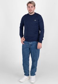 Granatowa koszula z długim rękawem i okrągłym dekoltem, z zielonym logo, w połączeniu z jasnoniebieskimi dżinsami i białymi trampkami, stojąca na jednolitym tle.