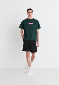 Camiseta de manga corta verde oscuro con un gráfico rosa. Combinada con pantalones cortos negros y zapatillas blancas en un modelo contra un fondo sencillo.