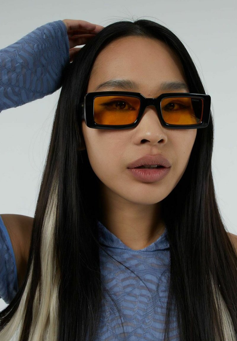 Des lunettes de soleil rectangulaires noires avec des verres orange, portées par une personne aux longs cheveux noirs, sur un fond neutre.