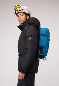 Zwarte geïsoleerde jas met een blauwe rugzak. Kenmerken zijn een hoge kraag, meerdere zakken en een helm met getinte goggles.