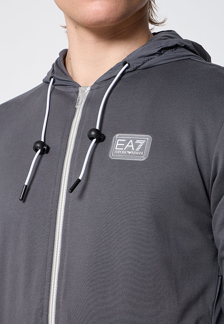 Felpa grigia con zip realizzata in tessuto morbido, dotata di cappuccio con cordini, un'etichetta con logo e dettagli della zip bianchi a contrasto.