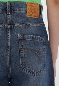 Blue denim jeans with a rear patch pocket featuring tan stitching. A leather label with an embossed logo is stitched on the waistband.