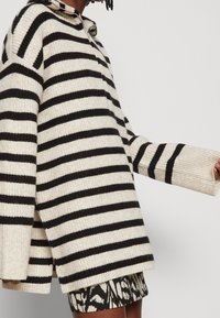 Personne portant un pull en maille beige avec des rayures horizontales noires, un col à fermeture éclair, et une jupe à motif noir et blanc.