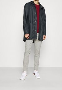 Personne portant un imperméable noir à capuche, une chemise rouge, un pantalon gris clair et des baskets blanches avec le swoosh rouge de Nike, se tenant sur un fond uni.