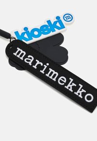 Svart stoffetikett med hvit brodert tekst "marimekko," ledsaget av en blå og hvit plastetikett med "kioski." Teksturerte designelementer er tydelige.
