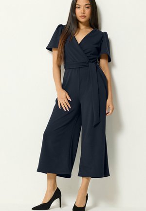 Mujer de pie que lleva un mono cruzado azul marino con mangas cortas y piernas anchas, combinado con zapatos negros de tacón alto.