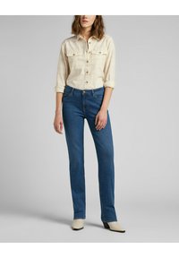 Camicia button-up beige chiaro con due tasche sul petto, abbinata a jeans blu a gamba dritta. Il modello indossa stivaletti bianchi con tacco.