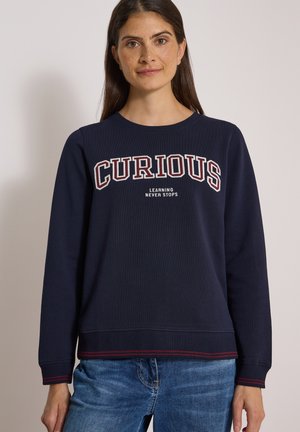 Frau trägt einen dunkelblauen Pullover mit den Worten "CURIOUS" und "LEARNING NEVER STOPS" und blaue Jeans, steht vor einem einfarbigen Hintergrund.