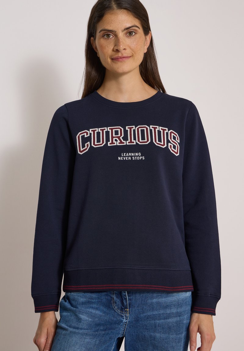 Femme portant un sweat-shirt bleu marine avec les mots "CURIOUS" et "LEARNING NEVER STOPS" et un jean bleu, debout devant un fond uni.