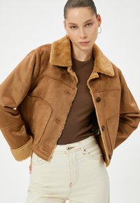 Veste en faux daim marron avec un col duveteux, des poches latérales et une fermeture à boutons ; assortie à un haut marron et à un jean de couleur claire.