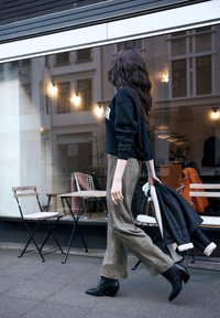 Sudadera negra, pantalones anchos color beige y botines negros con tacón. La modelo sostiene una chaqueta negra. Ambiente de café al aire libre. Elementos de diseño minimalistas.