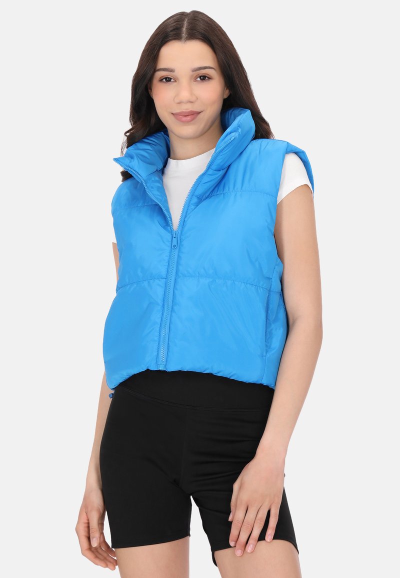 Vesta puffer croppată albastră, cu guler înalt, închidere cu fermoar și model matlasat. Puritată cu pantaloni scurți negri, peste o cămașă albă.