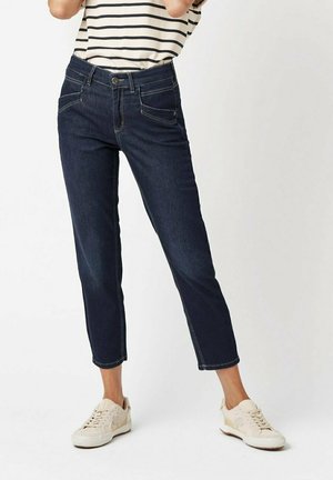 Dunkelblaue Jeans mit cropped Design, ausgestattet mit Vordertaschen, kontrastierenden Nähten und einem Knopfverschluss. Tragen mit hellen Sneakers.