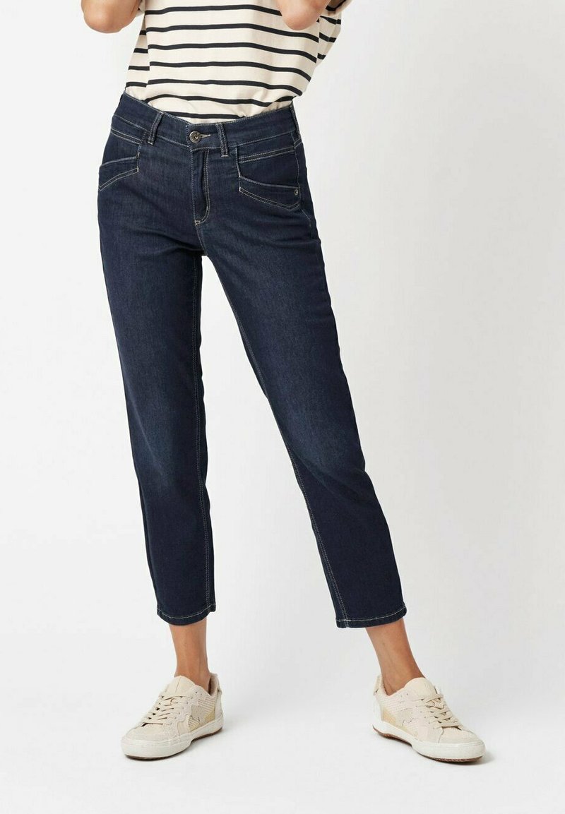 Dunkelblaue Jeans mit cropped Design, ausgestattet mit Vordertaschen, kontrastierenden Nähten und einem Knopfverschluss. Tragen mit hellen Sneakers.