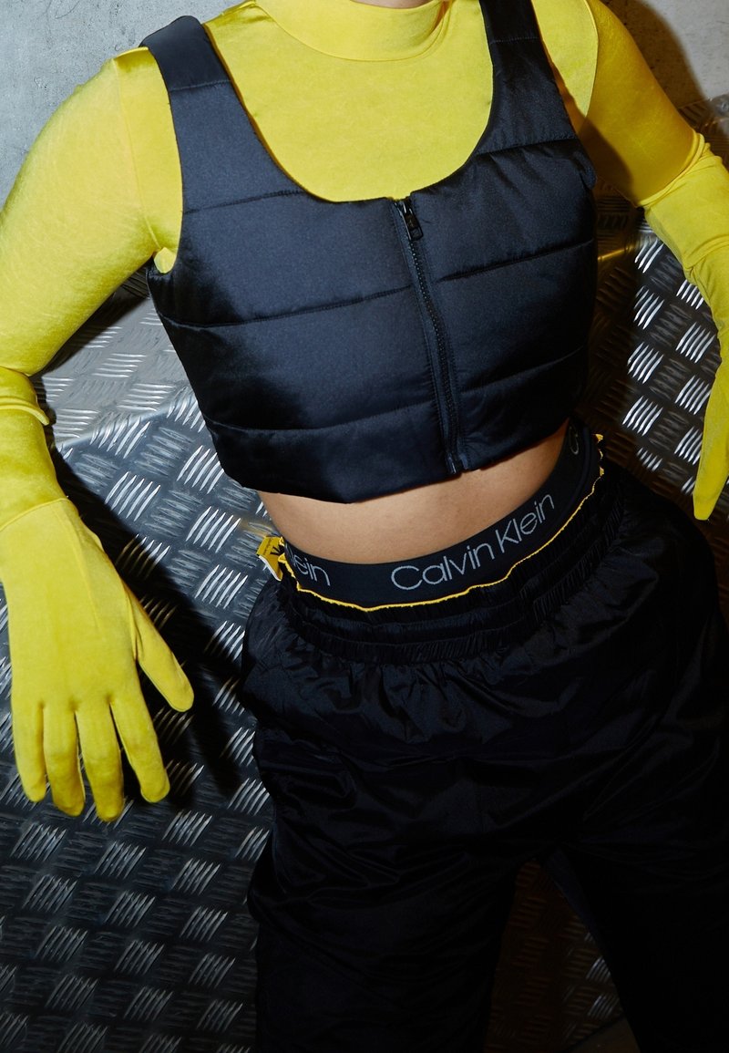 Personne vêtue d’un col roulé jaune avec des gants assortis, un gilet court noir, une ceinture Calvin Klein noire et un pantalon noir, assise sur un sol en métal.