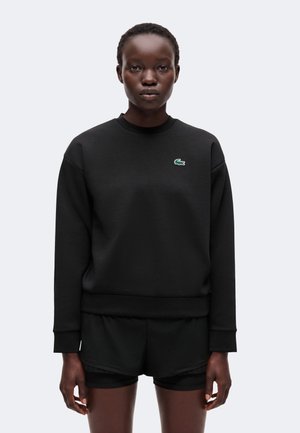 Persoon met een zwarte lange mouwen trui met een klein groen krokodillenlogo en zwarte korte broek tegen een effen lichte achtergrond.