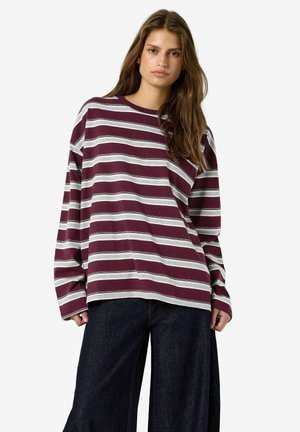 Femme aux longs cheveux bruns portant une chemise ample à manches longues rayée bordeaux et blanche et un pantalon large foncé, debout devant un fond blanc.