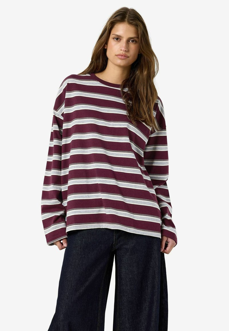Femme aux longs cheveux bruns portant une chemise ample à manches longues rayée bordeaux et blanche et un pantalon large foncé, debout devant un fond blanc.