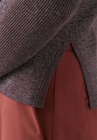 Närbild av en texturerad, mörkröd tröja med ribbat mönster och delad fåll, överlagd på ett rostfärgat tyg.