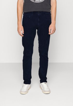 Mand iført mørkeblå slim-fit jeans, hvide sneakers og en sort grafisk t-shirt, stående mod en ensartet lys baggrund.
