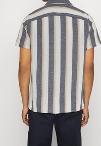 Koszula z krótkim rękawem w pionowe paseczki w kolorach granatowym, beżowym i białym; lekki materiał, luźny krój i kołnierzyk na guziki.
