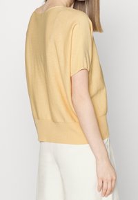 Krótki rękaw, lekki pastelowożółty sweter z okrągłym dekoltem, ściągaczem u dołu i luźnym krojem. W połączeniu z jasnymi spodniami.