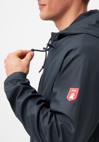 Námořnická dešťová bunda s kapucí, vybavená stahovací šňůrkou a červenou vyšitou nášivkou na rukávu. Hladký, voděodolný materiál.