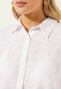 Camicia bianca ricamata con motivi floreali, apertura frontale con bottoni e bottoni perla. Presenta una texture morbida e un design classico del colletto.