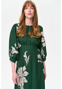 Zielona sukienka z gładkiej tkaniny, zdobiona dużymi białymi kwiatowymi wzorami, z marszczoną talią i bufkami. Prosty, okrągły dekolt.