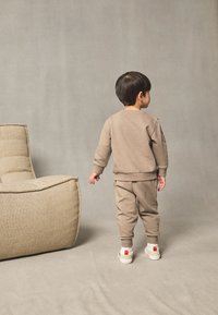 Bluză și pantaloni din bumbac de culoare bej deschis, cu croială lejeră. În prim-plan se vede un scaun texturat bej, pe fundal gri.