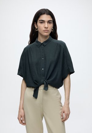 Femme aux longs cheveux foncés portant des boucles d'oreilles créoles dorées, une chemise boutonnée à manches courtes vert foncé nouée à la taille et un pantalon beige.