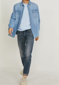 Ljust blå denimskjorta med tryckknappar, två bröstfickor; kombinerad med mörka jeans och beiga sneakers, som visar en avslappnad outfit.