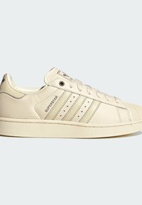Παλιοκίτρινο αθλητικό παπούτσι Adidas Superstar με διάτρητες τρεις ρίγες, λαστιχένια μύτη, κορδόνια και το κείμενο "SUPERSTAR" στο πλάι.