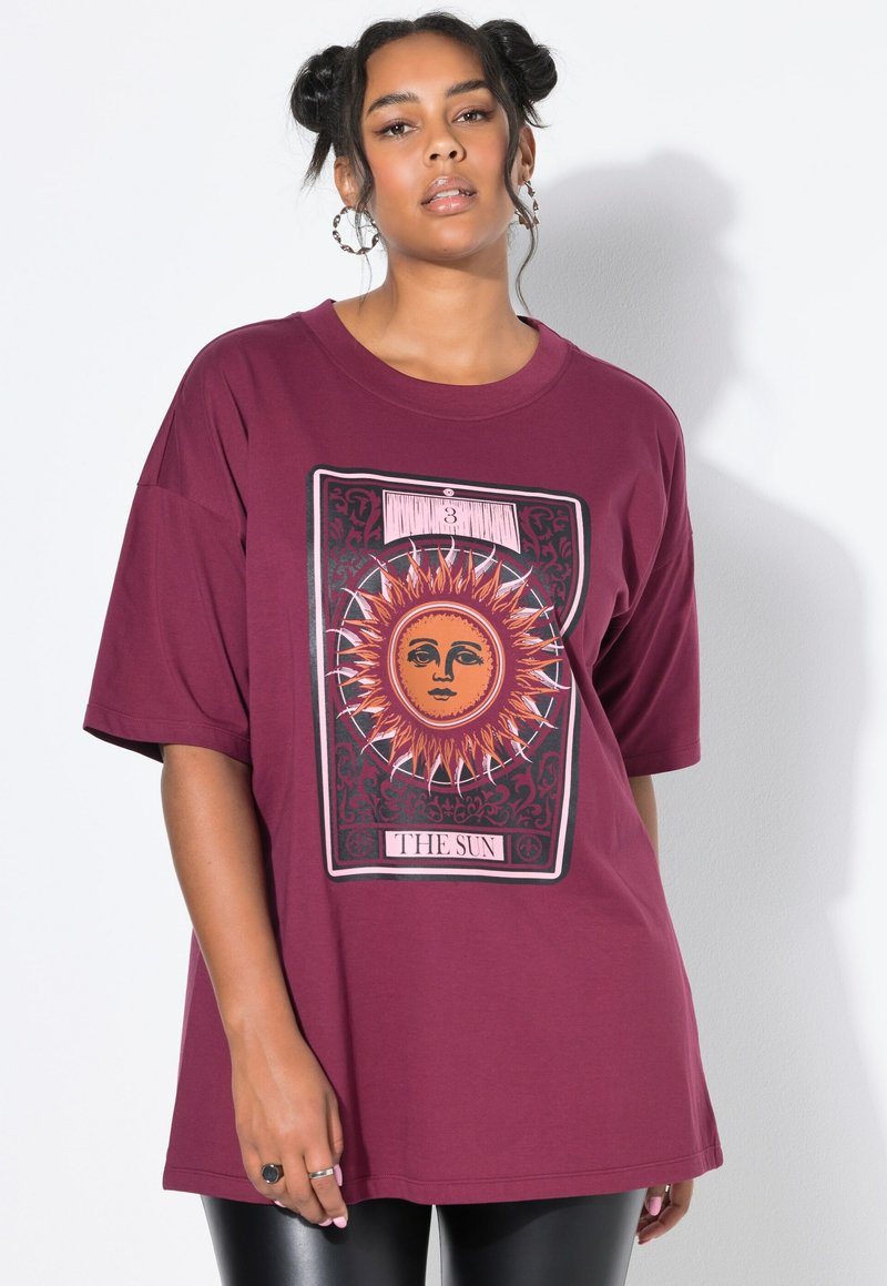Frau mit Space Buns Hairstyle, die große Ohrringe trägt und ein übergroßes T-Shirt in maroon mit dem Tarotkarten-Motiv "Die Sonne" trägt.