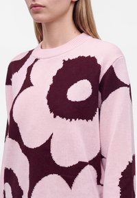 Femme portant un pull en tricot rose avec de grands motifs floraux abstraits violet foncé, montrant le haut du torse et le cou.