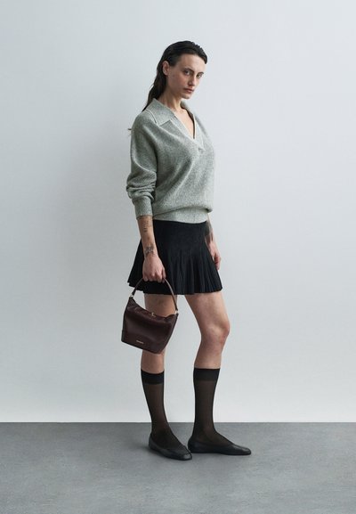 Grauer gestrickter Pullover mit V-Ausschnitt, schwarzer plissierter Rock, durchsichtige schwarze Socken und schwarze Ballerinas. Hält eine kleine Handtasche in dunklem Burgunderrot.