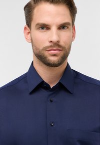 Maglietta blu navy scuro con colletto, tasca frontale e chiusura con bottoni. Tessuto liscio con una leggera lucentezza e design minimalista.