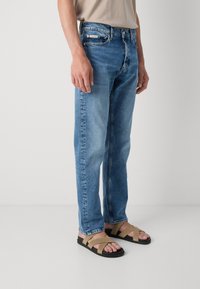 Modre denim hlače s ravnimi nogami, petimi žepi in svetlo pranjem, v kombinaciji z rjavimi in črnimi sandali z navzkrižnimi trakovi.