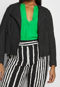 Giacca in pelle ecologica nera con chiusura a zip e accenti testurizzati sulle spalle, abbinata a una blusa verde e pantaloni a righe bianchi e neri.