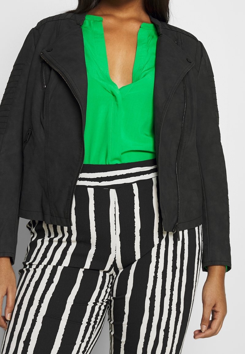 Giacca in pelle ecologica nera con chiusura a zip e accenti testurizzati sulle spalle, abbinata a una blusa verde e pantaloni a righe bianchi e neri.