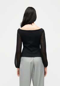 Γυναίκα με μακριά σκούρα μαλλιά που φοράει μαύρο τοπ έξω από τους ώμους με διάφανα μακριά μανίκια και ανοιχτό γκρι παντελόνι με ρίγες, εμφανιζόμενη από πίσω.