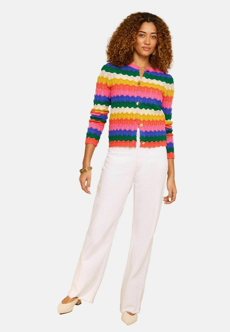 Lokkis juustega naine kannab mitmevärvilist triibulist kampsunit, valgeid laiade säärtega pükse, beežikaid kingi ja kuldseid käevõrusid valgel taustal.