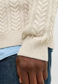 Cremfarbener Pulli mit Kabelstrickmuster, ausgestattet mit gerippten Bündchen und Saum. Kombiniert mit einem hellen Jeanshemd und dunklen Jeans, die Textur und Schichtung betonen.