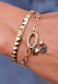 Pulsera en tono dorado que presenta una banda texturizada con tachuelas y una cadena con un eslabón circular, adornada con charms que incluyen una estrella, una perla y un disco decorativo.