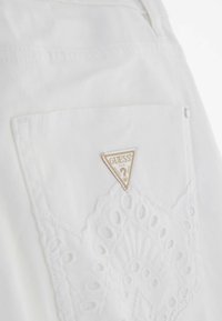 Vêtement blanc avec des détails en dentelle sur la poche, arborant une étiquette logo en forme de triangle. La texture est lisse avec des motifs brodés.