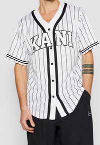 Maglietta da baseball bianca con strisce nere e logo "KANI". Presenta maniche corte, scollo a V e bottoni neri lungo la parte anteriore.