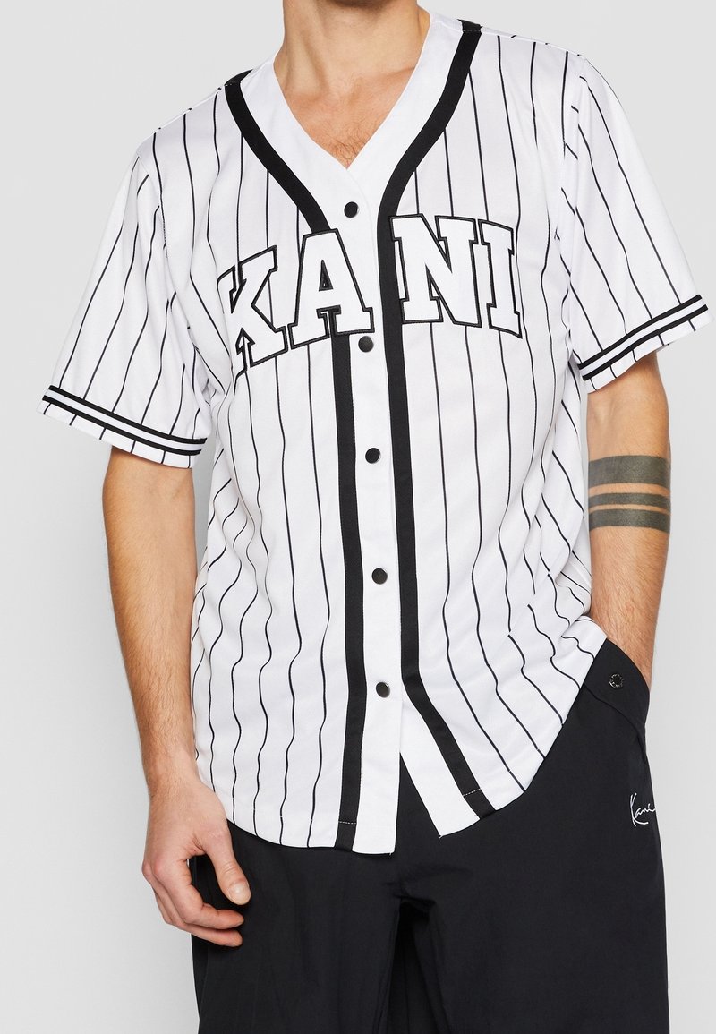 Maglietta da baseball bianca con strisce nere e logo "KANI". Presenta maniche corte, scollo a V e bottoni neri lungo la parte anteriore.