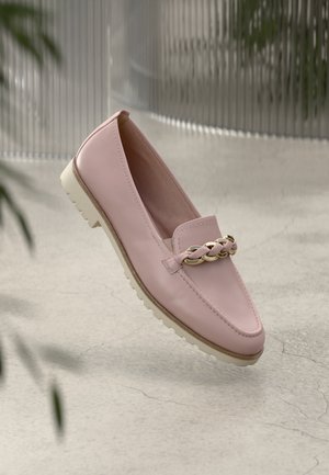 Pink leather loafer with braided gold and pink chain detail, white sole, floating above a cracked concrete floor with blurred plant edges.